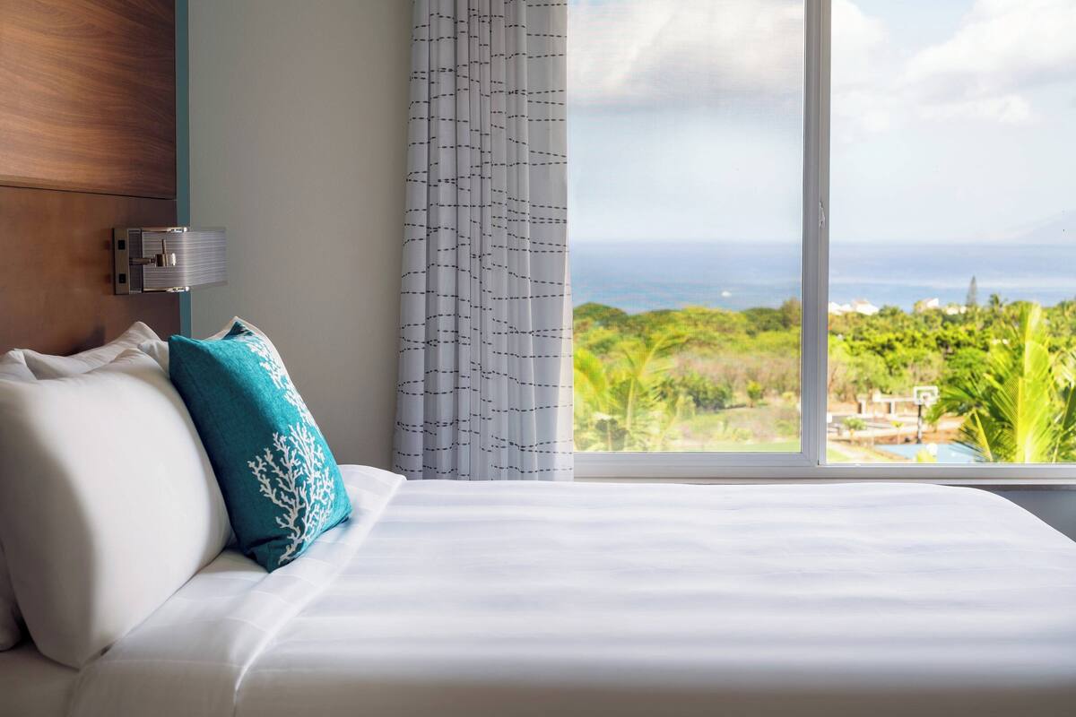 A hotel room with a bed, a view of the ocean, and a window with curtains.