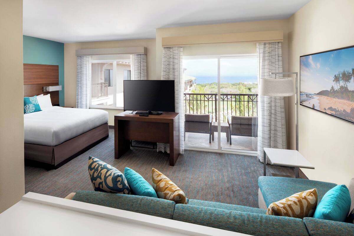A hotel room with a bed, a TV, a sofa, and a balcony with a view of the ocean.