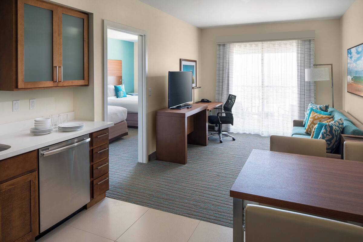 A hotel room with a kitchenette, a bed with teal bedding, a desk with a computer, a chair, and a sofa with patterned cushions.