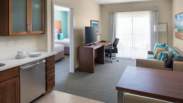 A hotel room with a kitchenette, a bed with teal bedding, a desk with a computer, a chair, and a sofa with patterned cushions.
