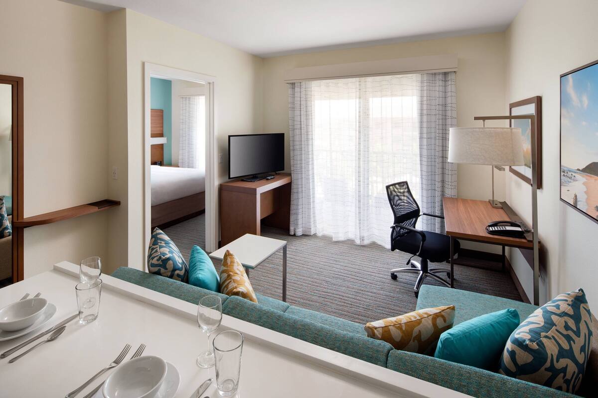 A hotel room with a bed, a TV, a desk, a chair, a dining table with cutlery, and a sofa with cushions.
