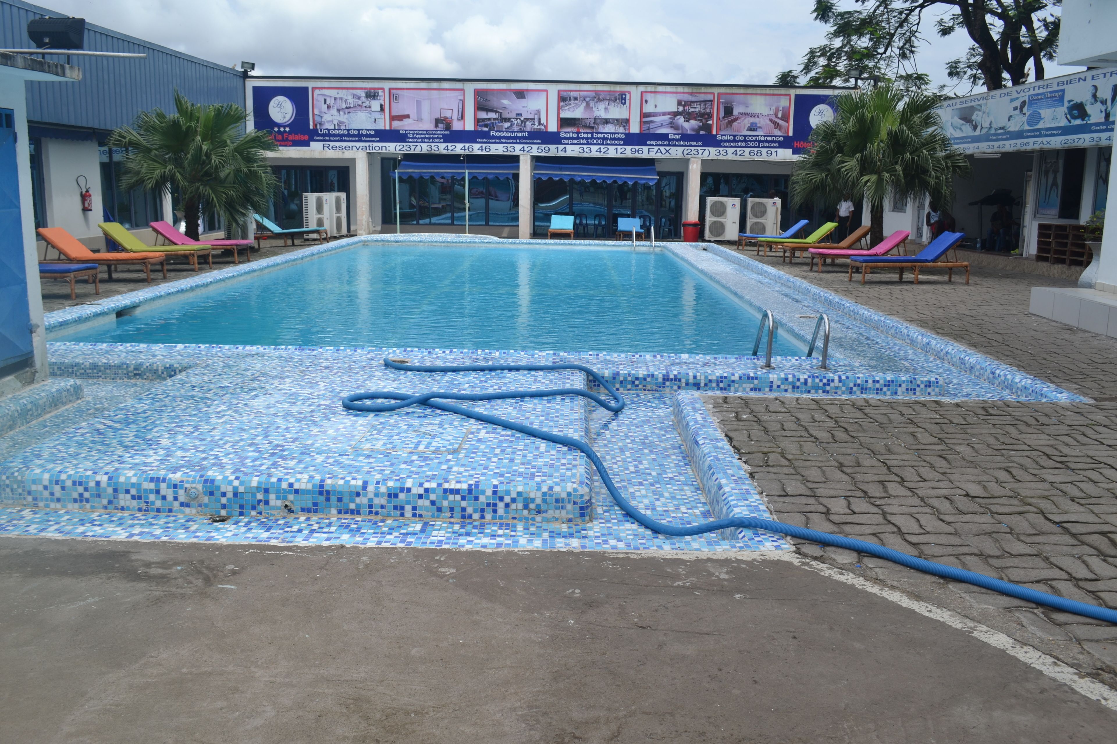 Una piscina con borde de azulejos, sillas de descanso y un edificio con señalización al fondo.