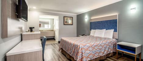 A hotel room with a bed, a desk, a chair, a TV, a bedside table, and a nightstand.