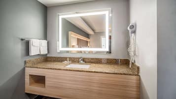 A modern bathroom with a granite countertop sink, a wooden vanity, a large mirror with integrated lighting, and a hairdryer mounted on the wall.