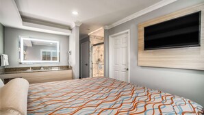Habitación de hotel con una cama grande, un televisor montado en la pared, un baño con espejo y lavamanos, y una zona de ducha.