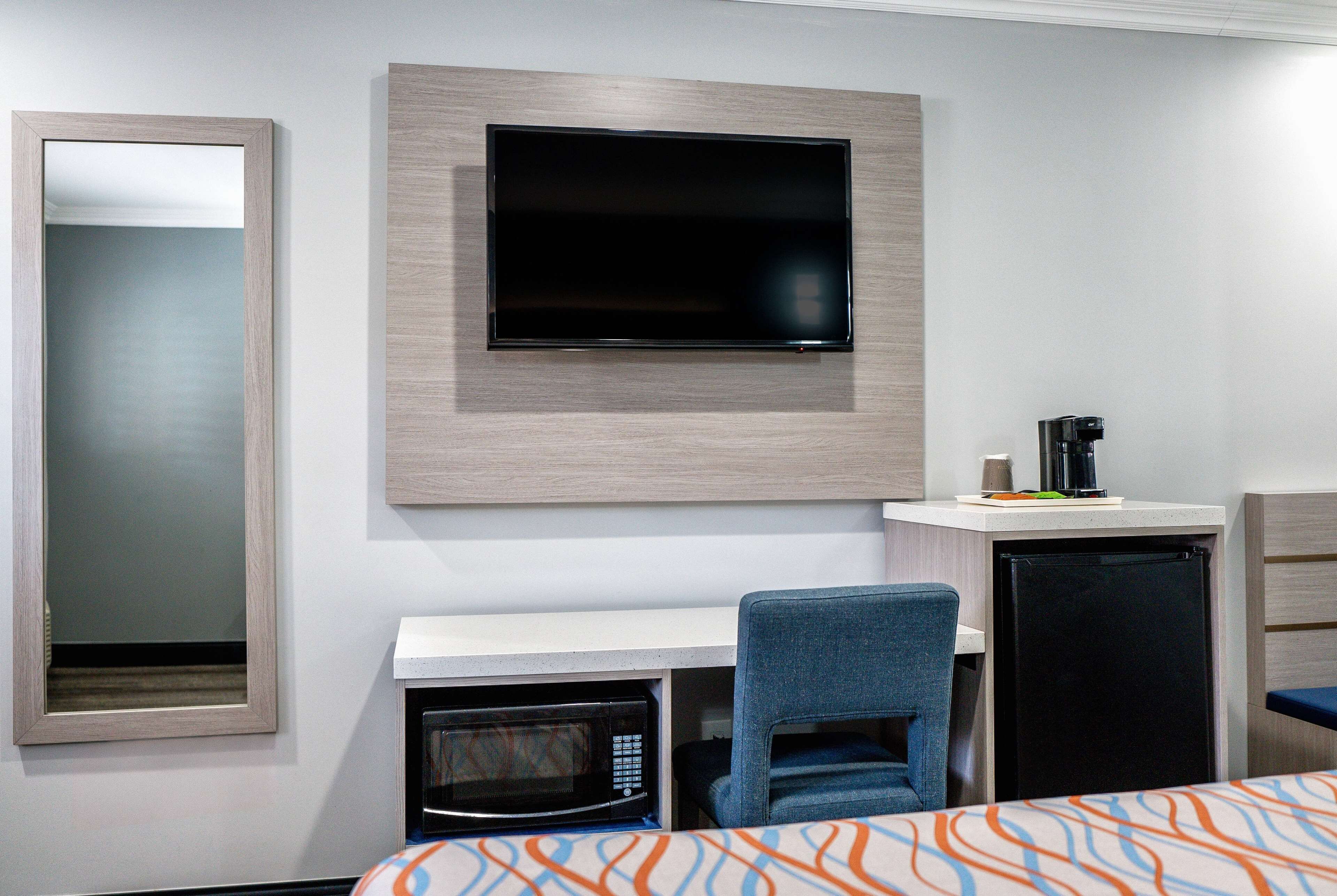 A hotel room with a flat-screen TV mounted on the wall, a microwave, a small refrigerator, a desk with a chair, and a bed with an orange and blue patterned bedspread.