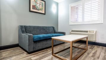 Une pièce avec un canapé gris, une table blanche, un tableau abstrait encadré, une fenêtre avec des stores et un climatiseur fixé au mur.