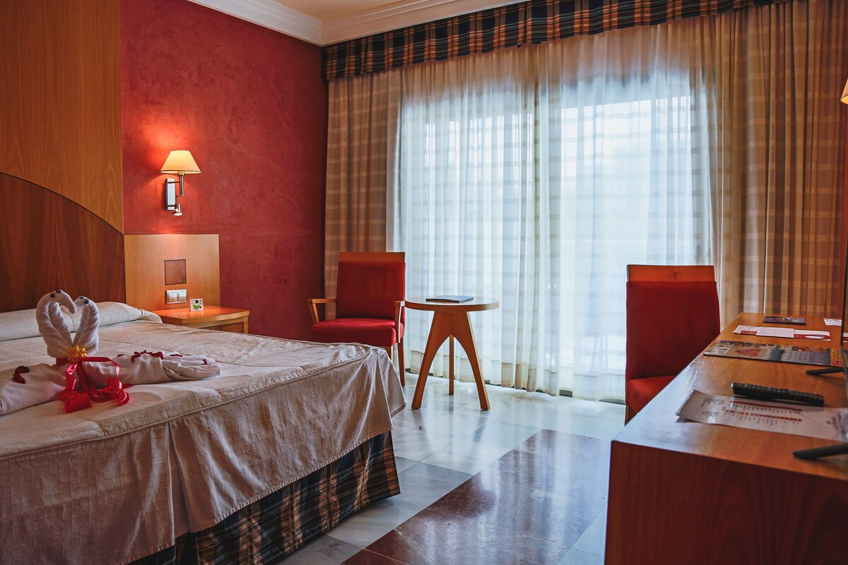 A hotel room with a bed, a desk, two chairs, a lamp, and a view of the outdoors through large curtains.