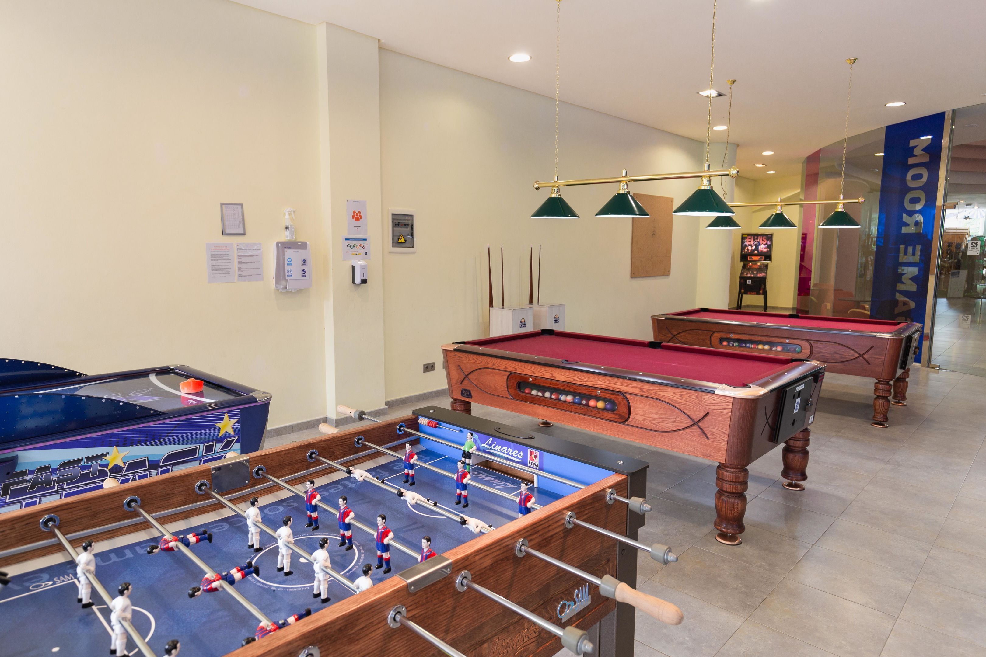 A pool table, a foosball table, and a billiards table in a room with a tiled floor and hanging pendant lights.