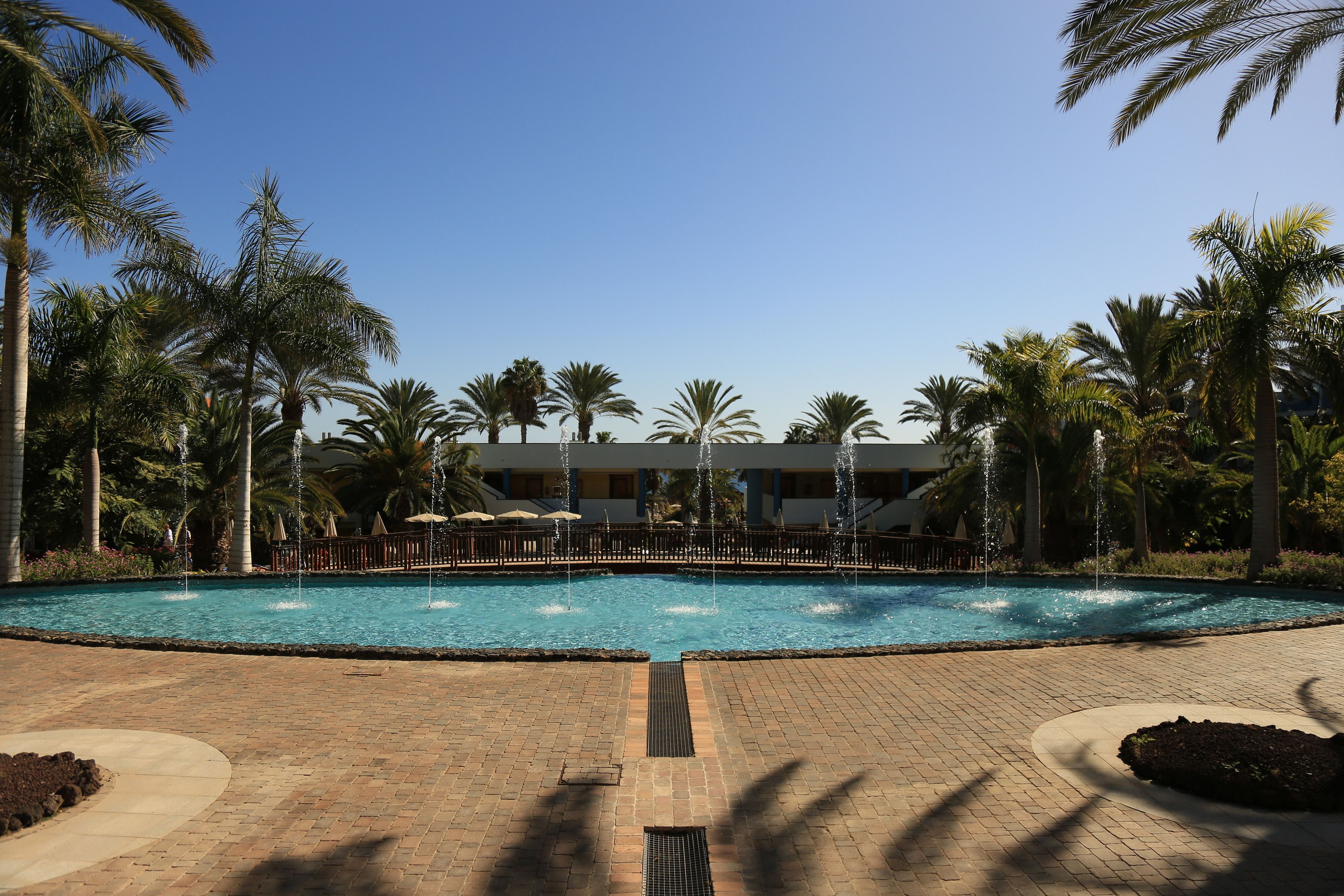 A swimming pool with water fountains, surrounded by palm trees and a brick pathway.