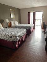 A hotel room with two beds, a desk, and a chair. There is a window with curtains and a door leading to an outdoor area.