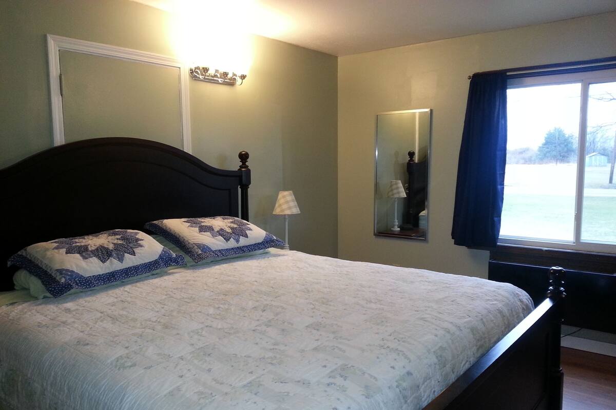 Un dormitorio con cama, mesita de noche, lámpara, espejo y ventana con cortinas.