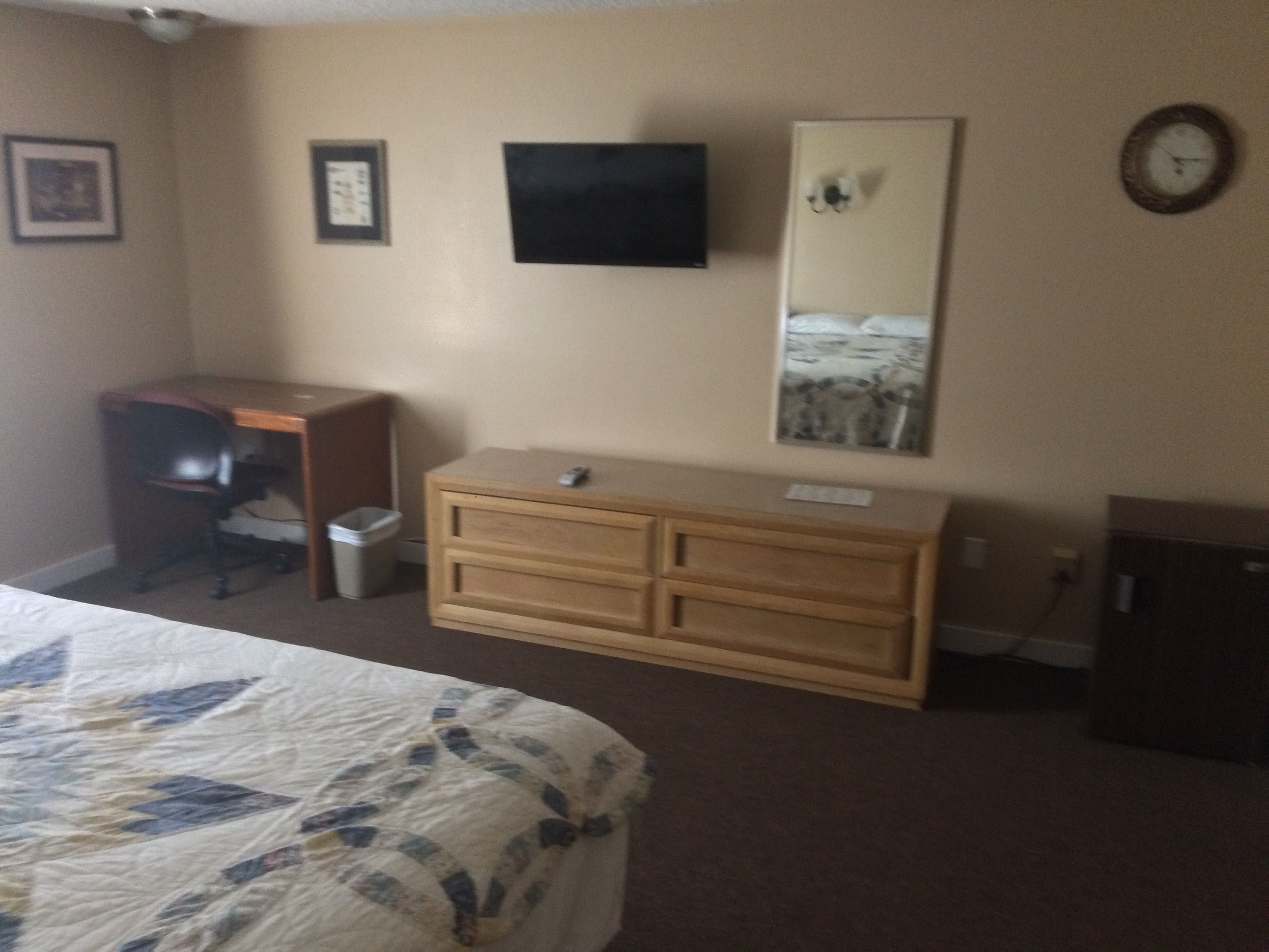 A hotel room with a bed, a desk, a dresser, a trash can, a television, and a clock on the wall.