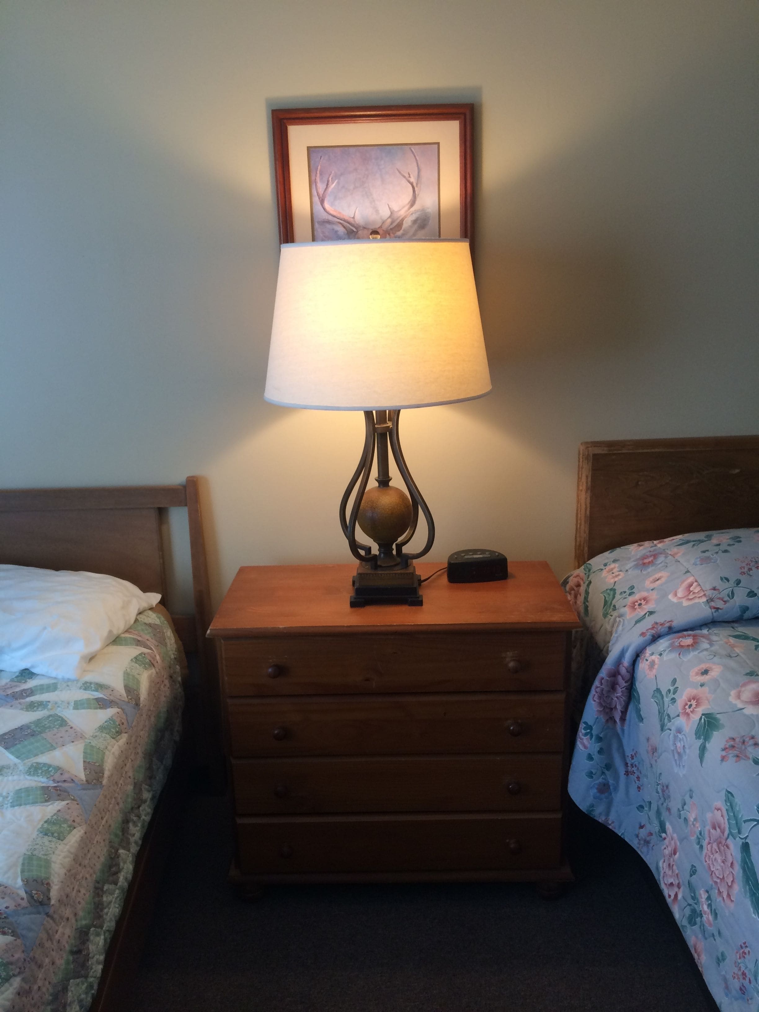 Et natbord i træ med en lampe, et indrammet billede over det og en seng med blomstret sengetøj til højre.