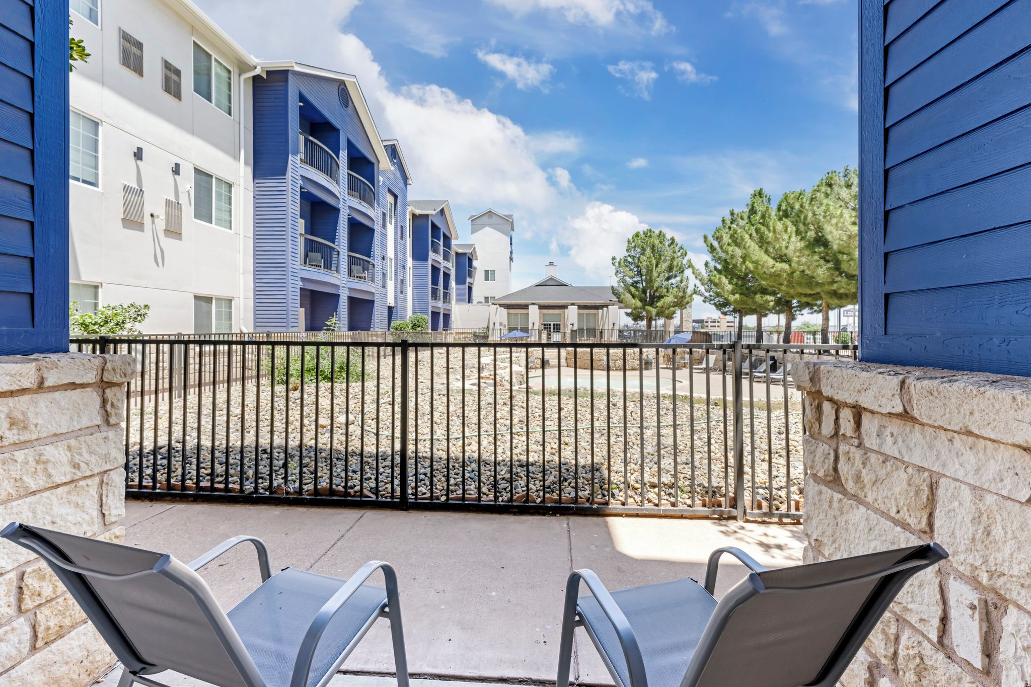 A balcony with two chairs, a stone wall, and a view of a multi-story building with balconies.