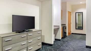 Habitación de hotel con televisor de pantalla plana sobre una cómoda de madera, un pequeño espacio de cocina con refrigerador y piso de baldosas.