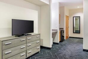 A hotel room with a flat-screen TV on a wooden dresser, a small kitchen area with a refrigerator, and a tiled floor.