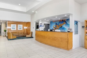 Un hall d'accueil d'hôtel avec un comptoir de réception en bois, un espace salon avec un canapé et des fauteuils, et un mur orné d'une inscription : « Bienvenue à Lubbock ».