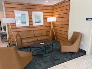 A lobby area with a brown leather sofa, a round coffee table, and two brown leather armchairs. There are two framed artworks on the wall and a sign indicating the location of restrooms. - Lubbock West Inn & Suites (Lubbock)
