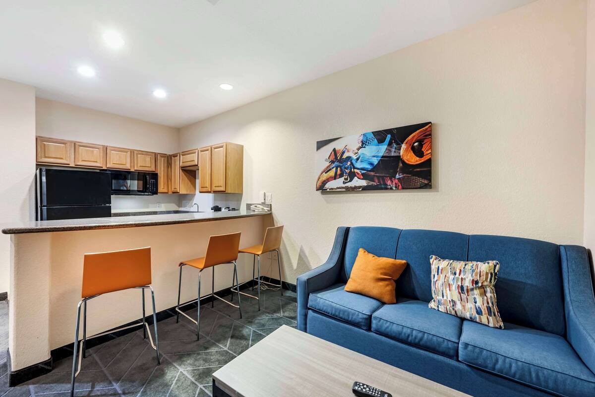 A modern kitchen with wooden cabinets, a black refrigerator, and a microwave. There is a blue sofa with a patterned pillow and a remote control on a wooden coffee table.