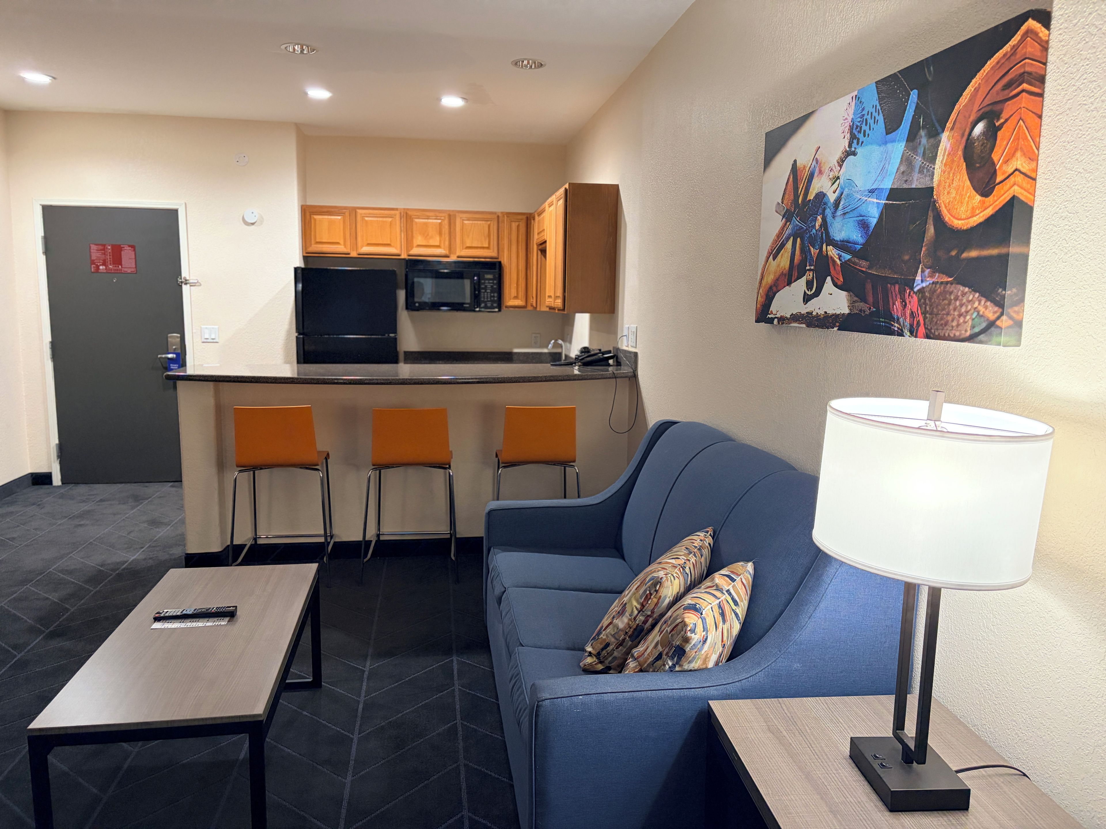 Une chambre d’hôtel moderne avec un canapé, une table, une lampe et, au fond, une kitchenette.