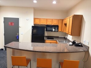 A compact kitchen with a refrigerator, microwave, and wooden cabinets. There are three orange chairs and a sink. - Lubbock West Inn & Suites (Lubbock)