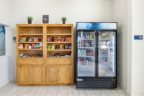 A kitchenette with a wooden cabinet stocked with snacks and a black refrigerator with a glass door. - Lubbock West Inn & Suites (Lubbock)