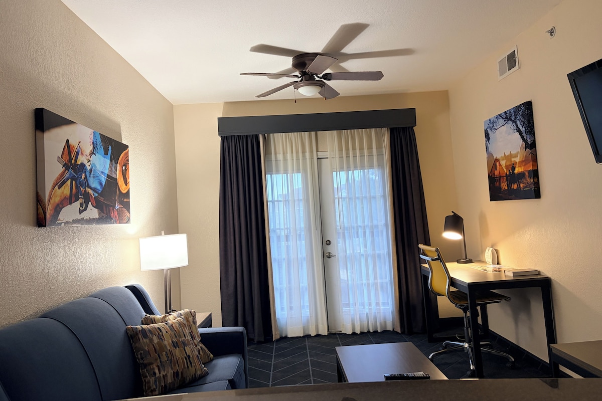 A hotel room with a ceiling fan, a desk with a chair, a couch, a window with curtains, and artwork on the walls.