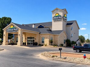 A Lubbock West Inn & Suites building with a stone facade, a prominent entrance, and a parking area with a fire hydrant.