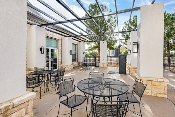 Un patio al aire libre con mesas y sillas redondas, una estructura de pérgola y una pared de piedra.