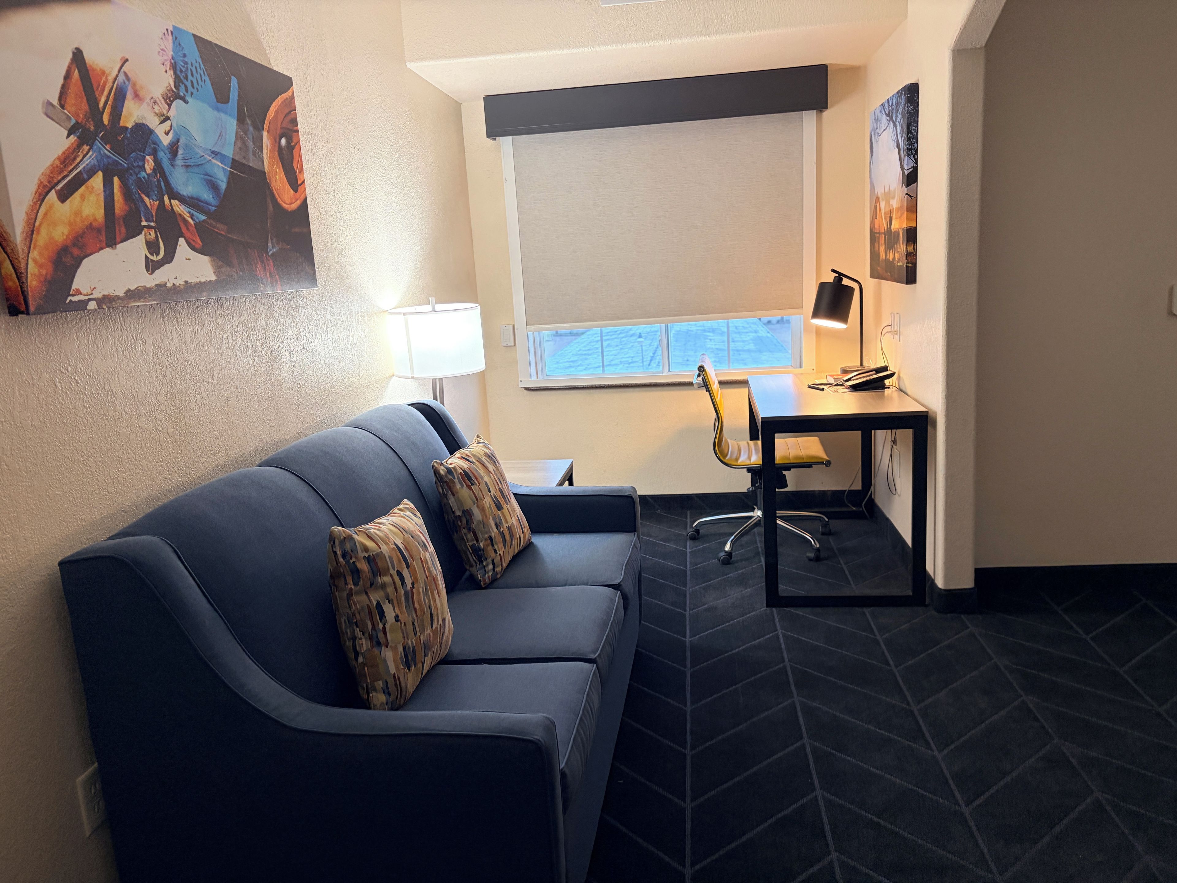 A modern room with a dark blue sofa, two patterned cushions, a desk with a chair, a lamp, and a large window.