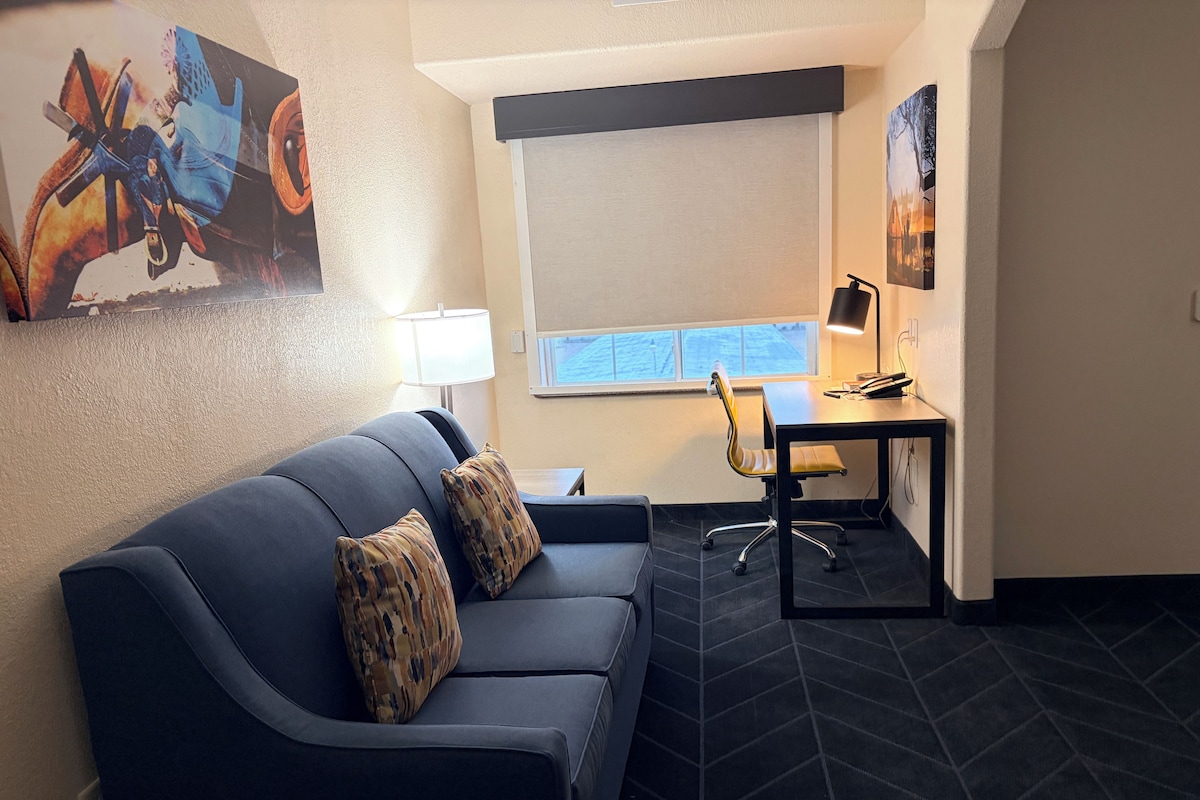 Une pièce moderne avec un canapé bleu foncé, deux coussins à motifs, un bureau avec une chaise, une lampe et une grande fenêtre.