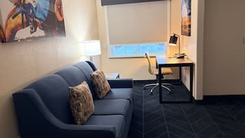 A modern room with a dark blue sofa, two patterned cushions, a desk with a chair, a lamp, and a large window.