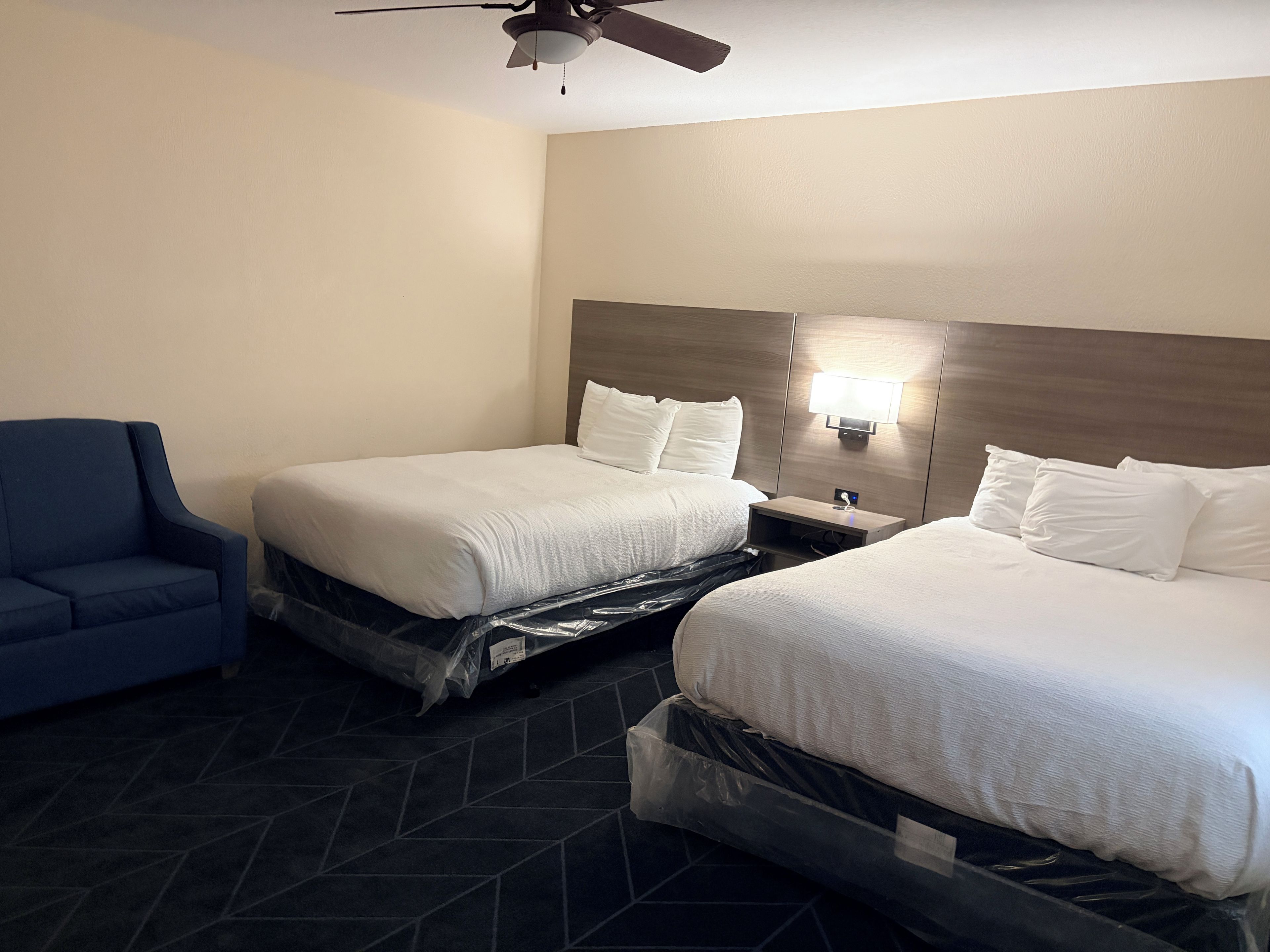 A hotel room with two beds, a blue armchair, a ceiling fan, and a nightstand with a lamp.