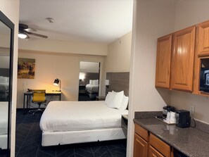 A hotel room with a bed, a desk with a chair, a kitchenette, and a ceiling fan. - Lubbock West Inn & Suites (Lubbock)