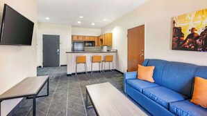A modern living room with a blue sofa, a flat-screen TV, and a kitchen area with wooden cabinets and a microwave. - Best Western Lubbock West Inn & Suites (Lubbock)
