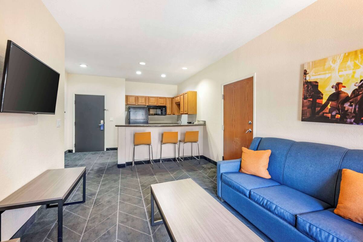 A modern living room with a blue sofa, a flat-screen TV, and a kitchen area with wooden cabinets and a microwave.
