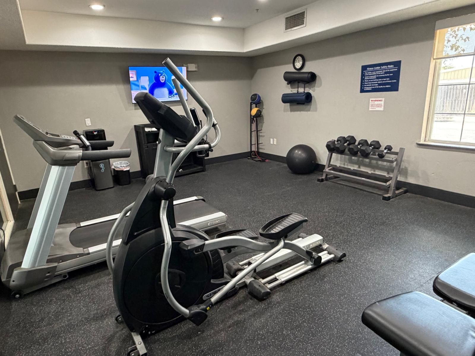 A gym with cardio equipment, dumbbells, and a TV screen.