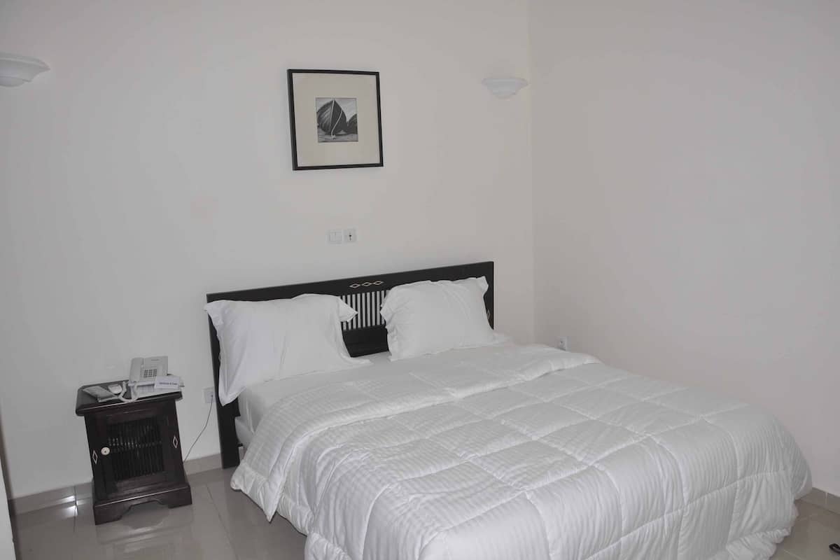 Una cama bien tendida con ropa blanca, una mesita de noche con un teléfono y una lámpara, y un cuadro en la pared.