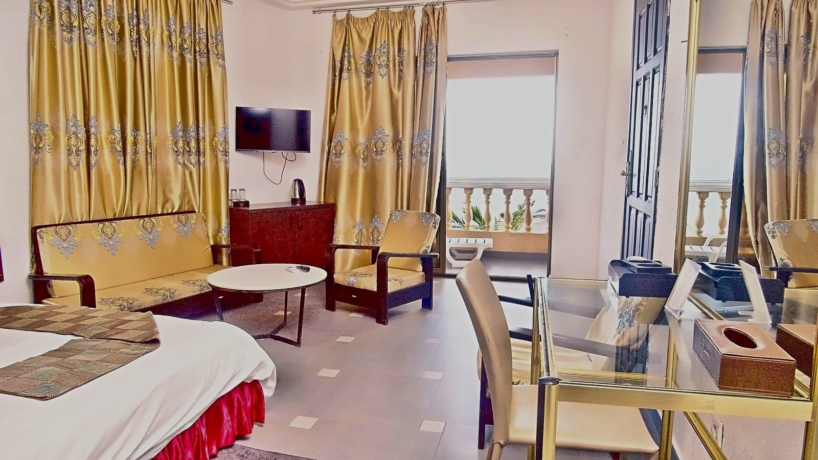 Une chambre d’hôtel avec un lit, une petite table ronde, un canapé, une télévision et un balcon doté d’une rambarde.