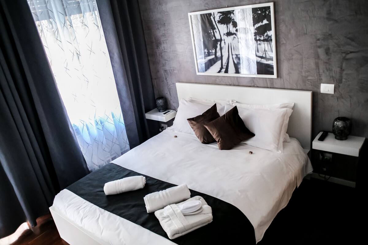 A neatly made bed with white and brown pillows, a black accent pillow, and two folded towels. There is a small bedside table with a vase and a remote control. A framed picture hangs on the wall above the bed.