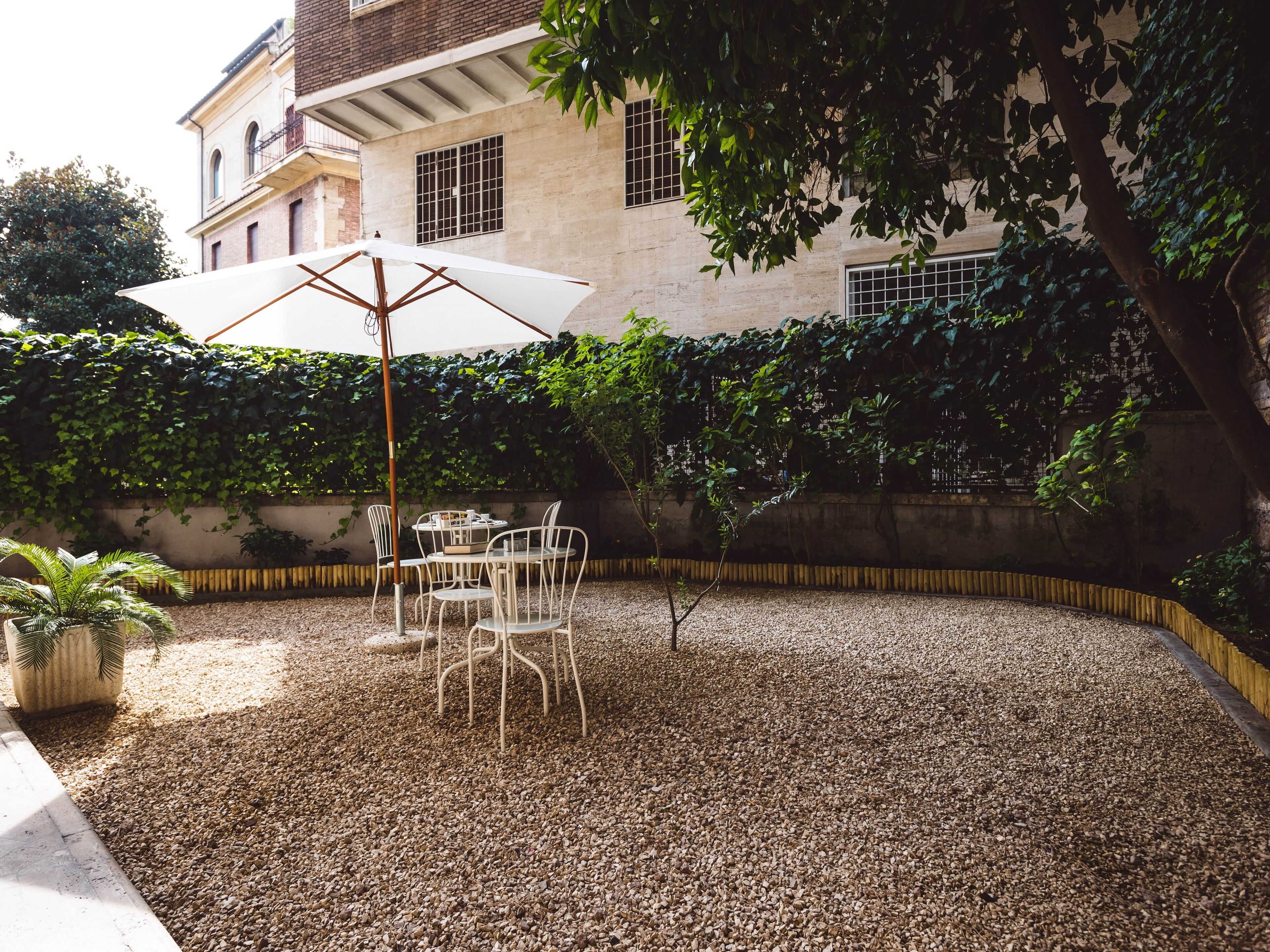 Un patio exterior con una mesa y sillas bajo un paraguas blanco, rodeado de grava y follaje.