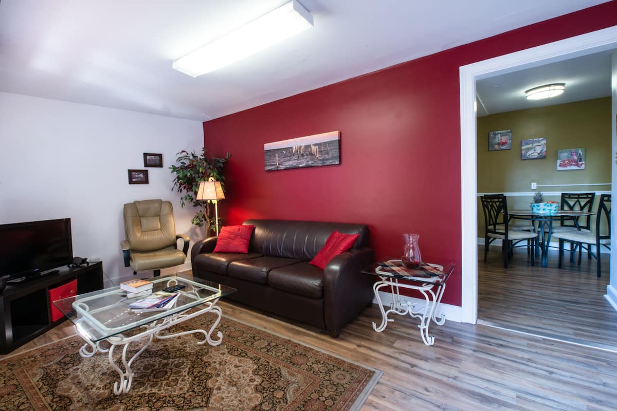 A living room with a brown leather sofa, a glass coffee table, a red wall, and a dining area visible through an open doorway.