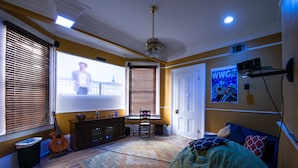 Una habitación con un ventanal grande, un ventilador de techo, una cama, una guitarra y una pantalla de proyector.