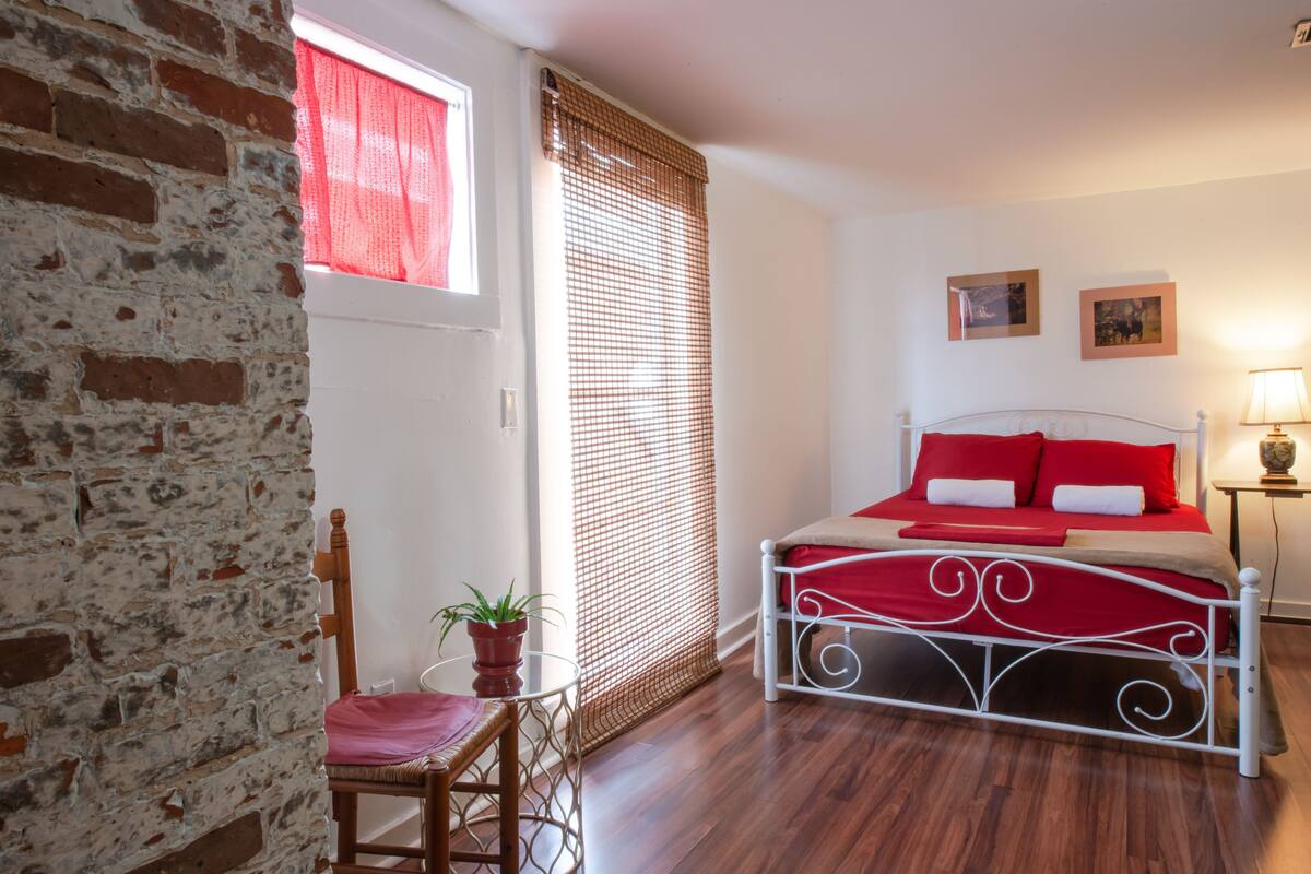 A bedroom with a brick wall, a window with red curtains, a bed with white metal frame and red bedding, a wooden chair, and a small table with a potted plant.