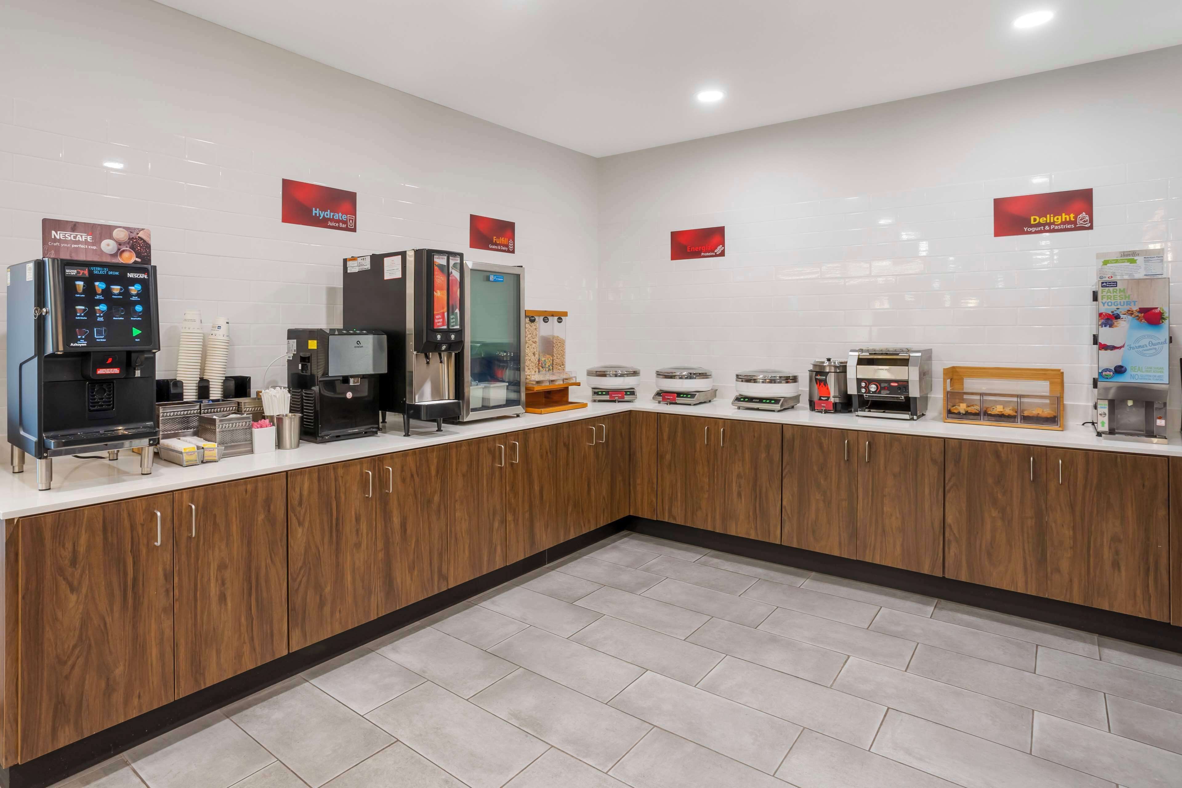 A self-service breakfast area with coffee machines, a yogurt dispenser, and a refrigerated display.