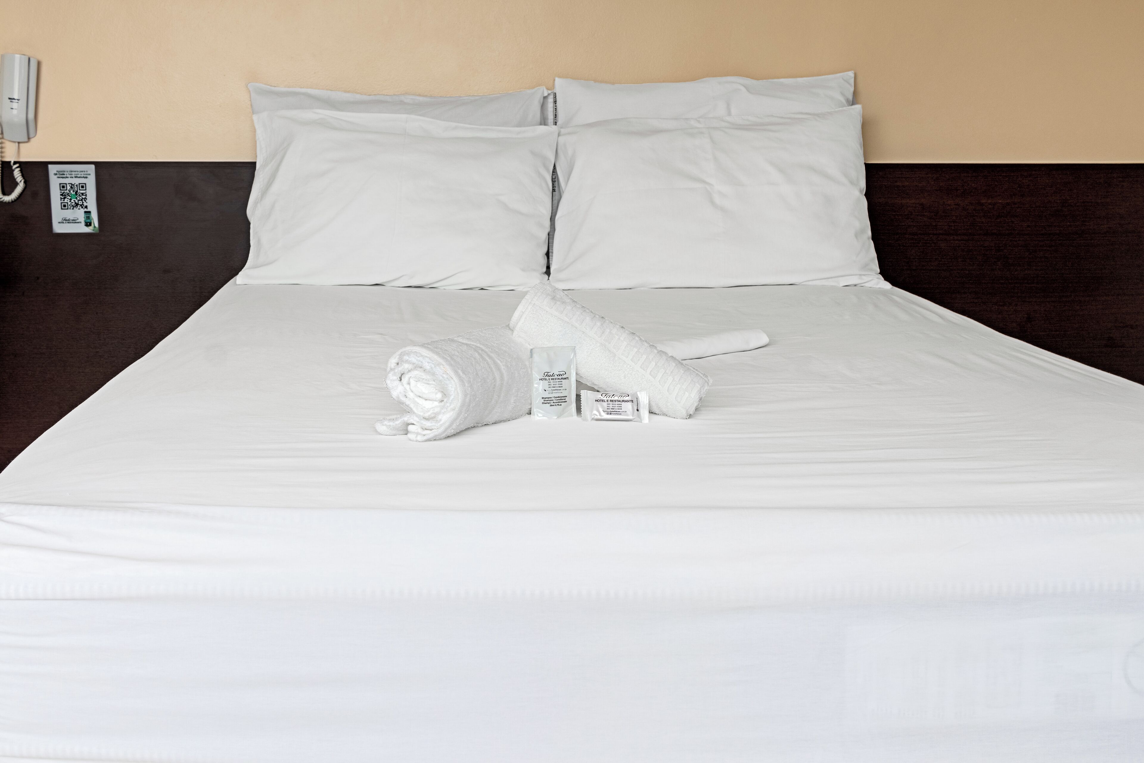 Una cama bien hecha con sábanas blancas, una toalla enrollada y artículos de aseo.