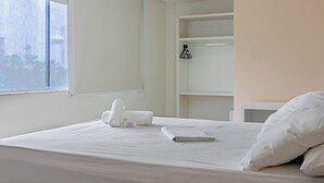 A neatly made bed with white linens and pillows, a window with a view of buildings, and a white wall with a shelf holding a black hanger.