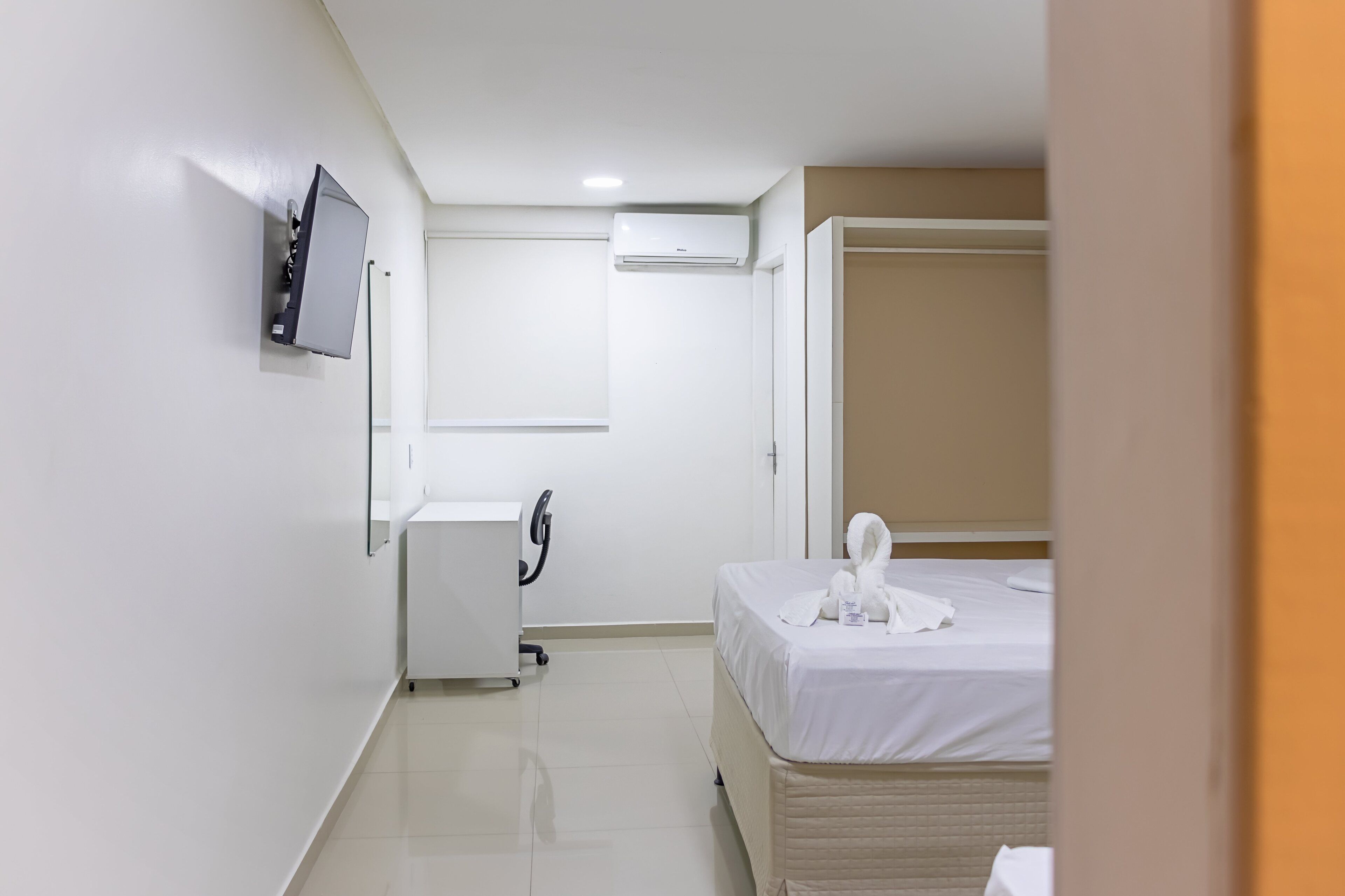 A hotel room with a bed, a desk with a chair, a flat-screen TV mounted on the wall, and an air conditioning unit.