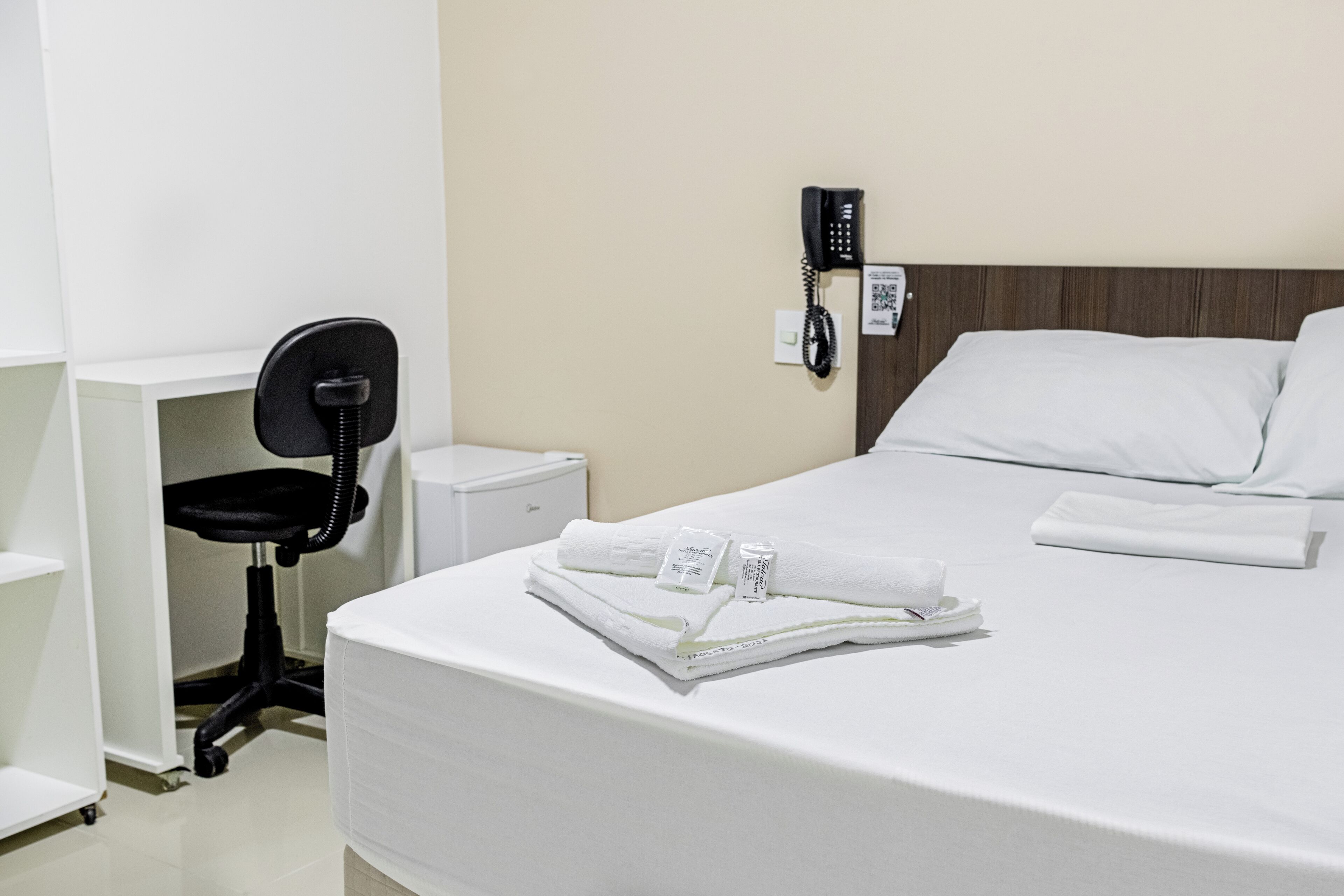 A hotel room with a neatly made bed, a desk with a chair, a telephone, and a small bedside table.